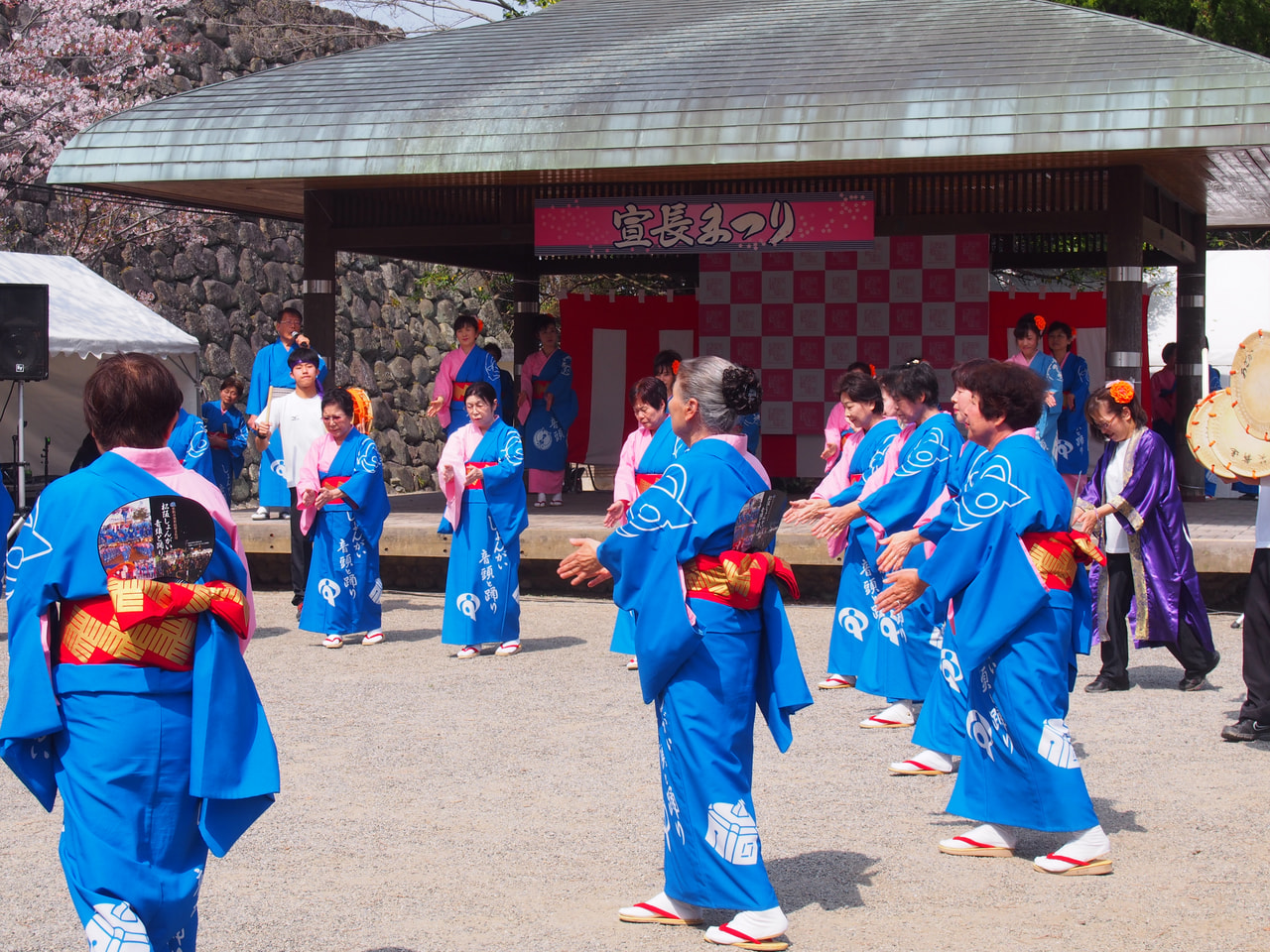 Women dressed in yukata are dancing in a circle facing the circle's center in front of a wooden stage.